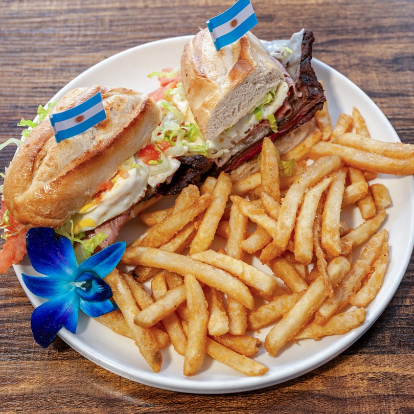 Two Argentine sandwiches with steak, egg, and vegetables, served with fries and a blue orchid.
