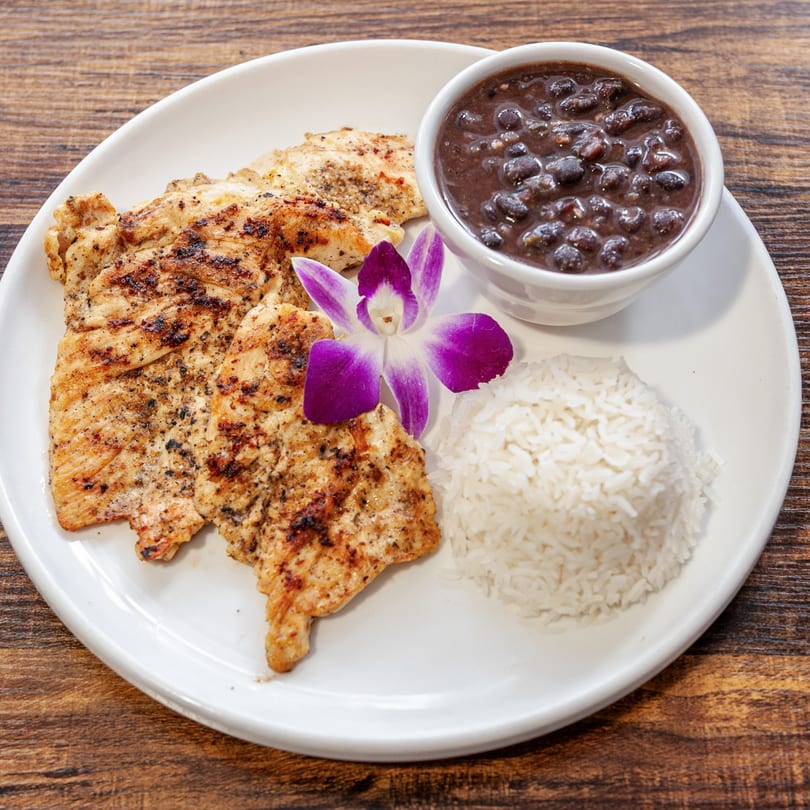 Grilled chicken, white rice, and black beans on a white plate, garnished with a purple orchid.