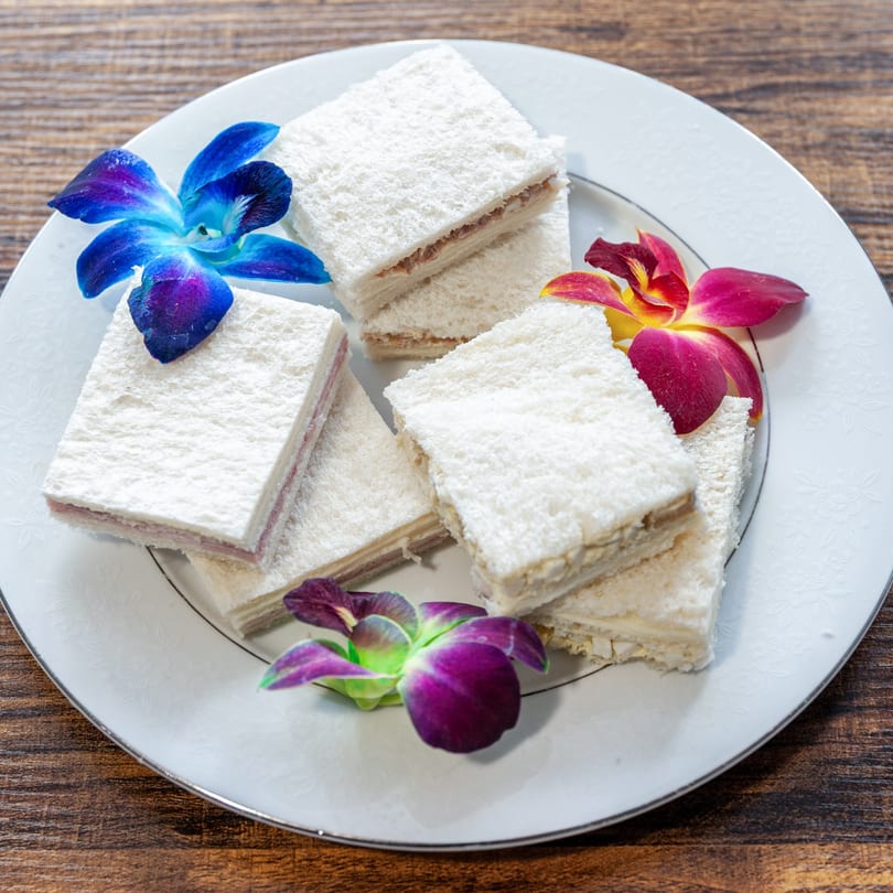 Small, crustless sandwiches with various fillings, garnished with vibrant orchids on a white plate.