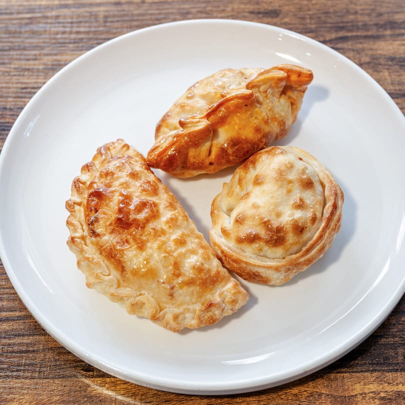Three golden-brown Dulce Bakery empanadas with crimped edges are arranged on a white plate.
